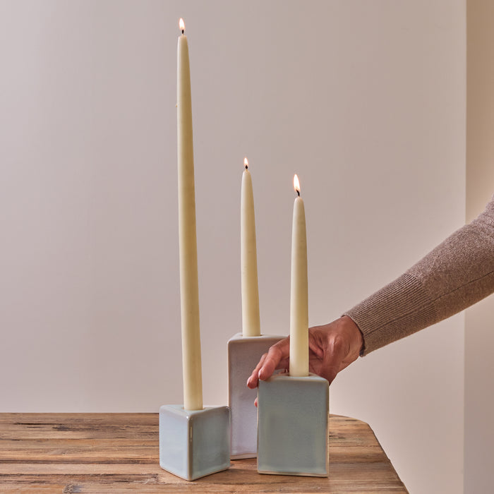 Three cream-colored taper candles in gray ceramic cube holders on a wooden surface.