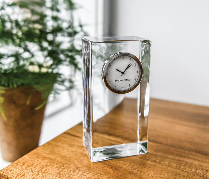 Tall rectangular clear glass clock with round metal-rimmed dial showing time, displayed on wooden surface next to potted plan