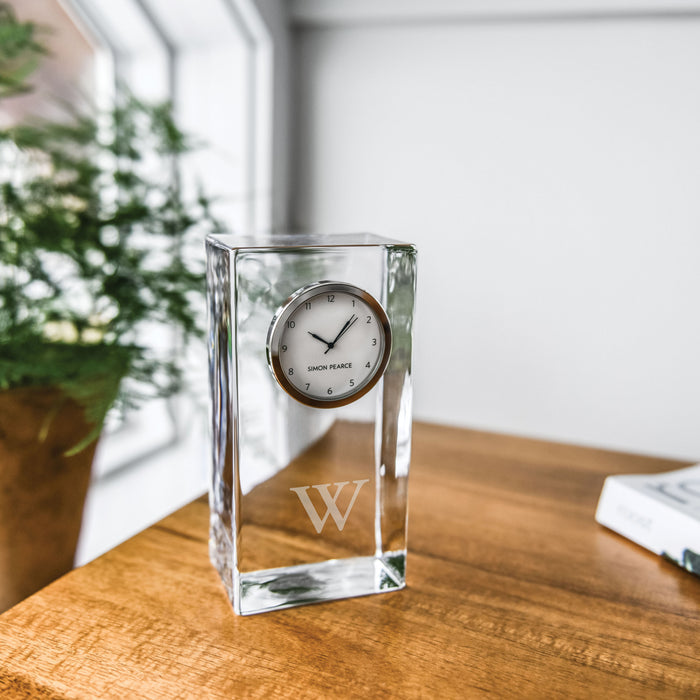 Clear glass rectangular clock with circular dial and etched "W" on front, displayed on wooden surface.