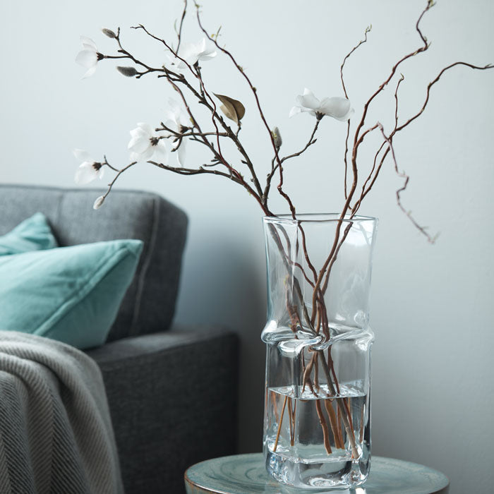 Clear glass vase with twisted cylindrical sections holding branches with white flowers and brown stems.