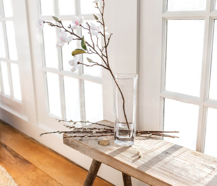 Clear glass cylindrical vase with brown branches and pink flowers on a wooden table by a window.