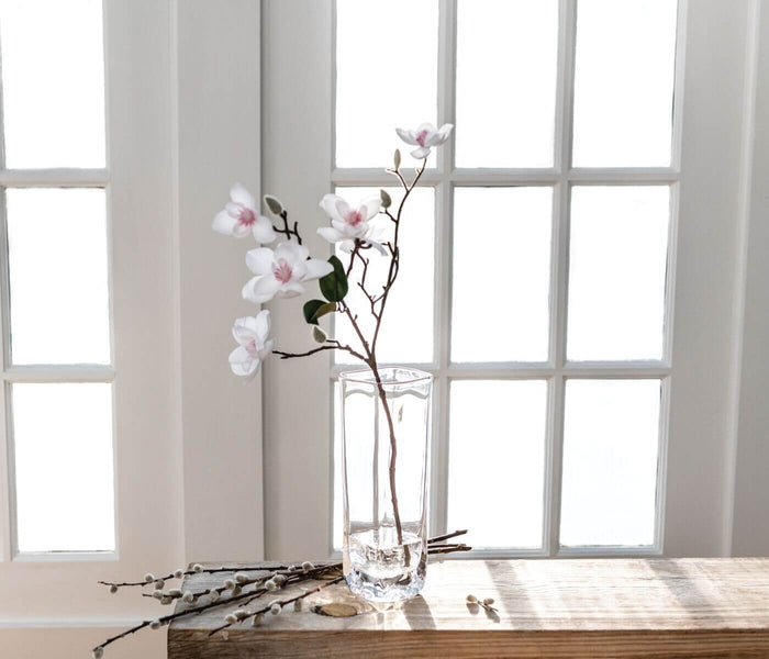 Clear glass cylindrical vase with pink and white orchid stems and branches arranged inside, displayed on wooden surface.