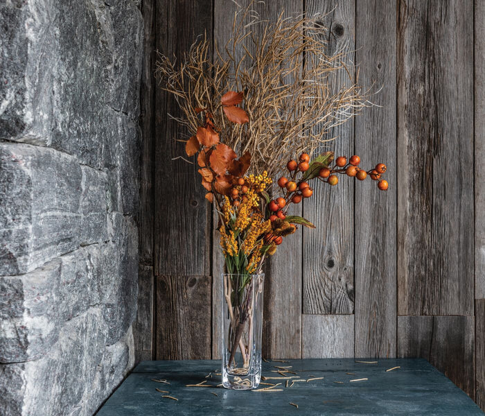 Clear glass cylindrical vase filled with dried grasses, orange berries, and rust-colored leaves.