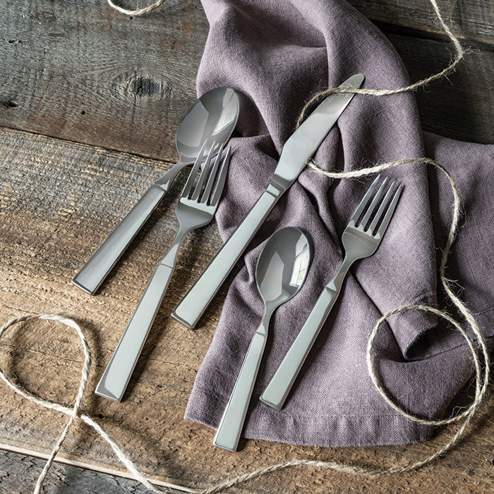 Five stainless steel flatware pieces with tapered handles arranged on a lavender napkin with twine on a wooden surface.