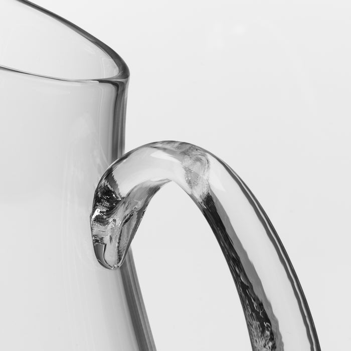 Clear glass pitcher with curved handle and spout, shown in close-up detail against white background.