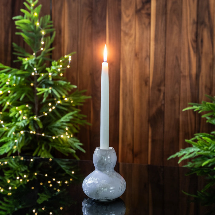 Blue-gray pottery candlestick with bulbous base holding a lit white taper candle, surrounded by evergreen branches.