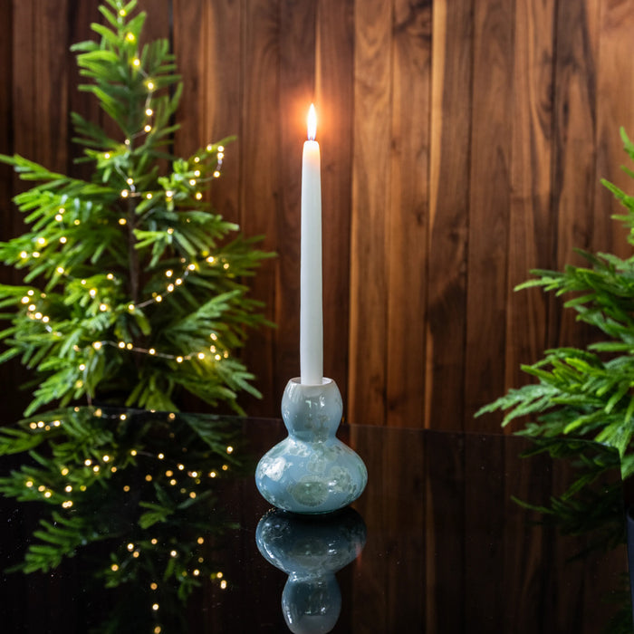 Blue pottery candlestick with bulbous base holding white lit candle, flanked by evergreen branches with small lights.
