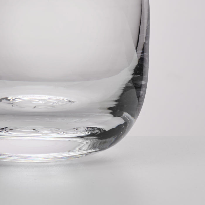 A close-up studio shot of a clear glass tumbler, showing the curved base and a portion of the transparent body against a light gray background.