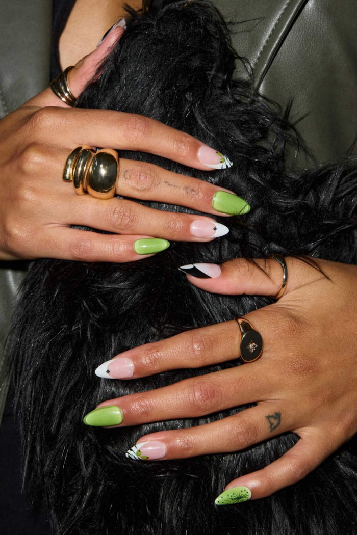 Short almond press-on nails: green, French tips, abstract lines, leaf designs. Adorned with gold rings.